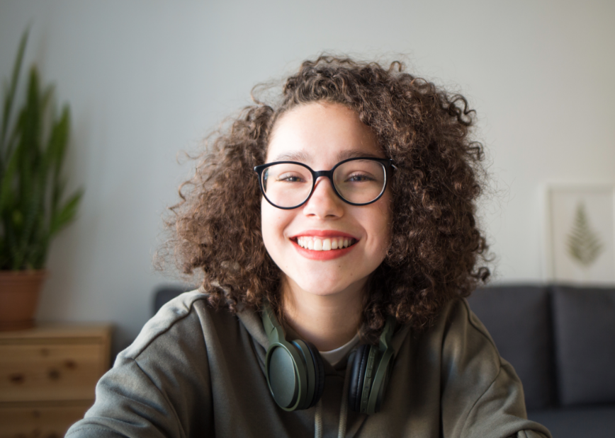 Teen girl with glasses, curly hair, red lipstick, headphones, and a jacket smiling